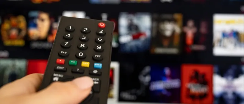 A hand holds a black TV remote control in the foreground, pointing towards a television screen that displays a blurred grid of movie and show thumbnails from a streaming service.